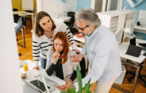 Trois collègues discutant autour d’un ordinateur portable dans un bureau lumineux, collaborant sur un projet ou une tâche administrative.